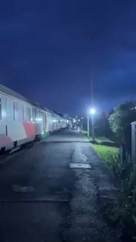 The video shows a train station at night. A train is stopped alongside the platform. The platform is wet and illuminated by overhead lights. A few people are visible in the distance. The sky is a deep blue. Someone says "Yo, we just got stuck in here. Somebody just pulled the emergency leach."
