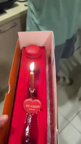 A hand holds a rectangular cake box containing a narrow red cake. On the cake is a lit candle, a red macaron, and a heart-shaped tag. The cake is then brought near an older bald man sitting at a table with his hands clasped. The table has a patterned table cloth. People out of frame are singing. A walker is visible in the background.