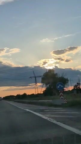 The video shows a view from a moving vehicle on a highway at sunset. Tall electricity pylons with wires are visible in the distance. The sky is a mix of blue and orange hues with scattered clouds. The highway has white markings and a guardrail on the side. A truck is visible in the distance on the highway.