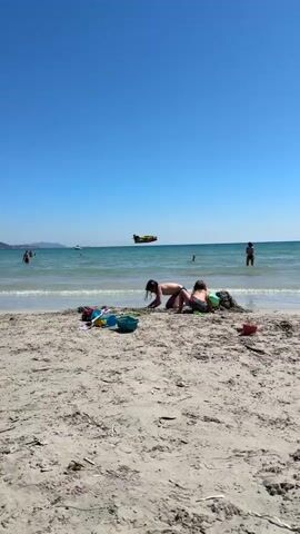The video captures a bright, sunny beach day in La Ciotat, France. The foreground shows a sandy beach where two children are seated, engrossed in playing with sand, surrounded by colorful beach toys and a black bag.

Beyond the beach, the calm, clear turquoise waters of the sea extend under a cloudless blue sky. Several people are visible in the water, some wading close to the shore, while others are swimming further out. Distant boats and a faint coastline can be seen on the horizon.

A notable event unfolds as a yellow seaplane, likely a Canadair firefighting aircraft, flies very low over the water, moving from left to right across the entire frame. Its flight path suggests a water scooping maneuver, common for such planes during training or in response to wildfires. The strong sunlight indicates it is likely midday or early afternoon. The scene combines the leisurely atmosphere of a beach outing with the dynamic presence of the specialized aircraft. No visible text is present.