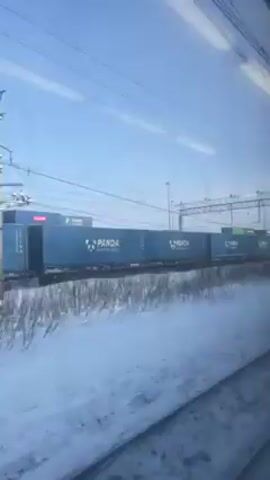 The video shows a view from a train window, looking out at a passing freight train. The freight train is carrying shipping containers, many of which are blue with the word "PANDA" written on them in large, white letters, along with a white panda logo. Some containers are green. The ground is covered in snow. There are power lines overhead. The sky is blue and the weather appears sunny.