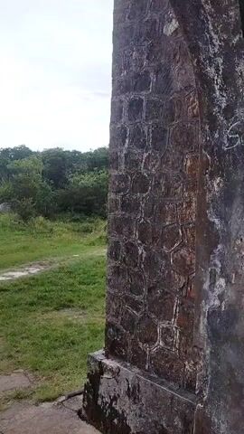 The video begins by showing a close-up of a stone archway and wall, transitioning to a blurry view of an interior with water damage and graffiti. It then shows a stone altar or table in a room with white walls and a concrete floor, before panning to the left to reveal an empty stone structure with greenery in the background. The video ends with a view of the altar from a different angle, showcasing its simple, rough-hewn design.