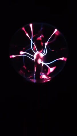 A plasma ball is shown against a black background. The plasma ball emits pink and blue tendrils of light that flicker and move within the glass sphere. The camera is positioned in front of the plasma ball, capturing the dynamic display of light.
