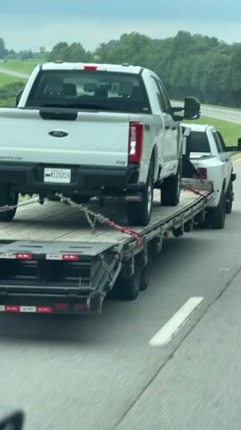 A white Ford Super Duty pickup truck is being transported on a flatbed trailer on a highway. The trailer is being pulled by another white truck, only partially visible, which is likely also a pickup truck. The Ford Super Duty is secured to the trailer with chains and straps. The video appears to be shot from another vehicle on the highway, looking towards the back of the trailer. The sky is overcast, and there are trees and rolling hills in the background. Other vehicles are visible in the distance on the highway.