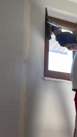 A person, wearing a white t-shirt, red pants, and blue gloves, is applying white plaster or wall filler to an interior wall. They are using a large trowel and a smaller putty knife to scoop and spread the material. The work is being done near a window with a brown frame, through which daylight is visible. Tattoos are visible on the person's right arm. Part of the wall already has a smooth, white finish, while another section is being worked on.