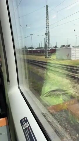 The video is shot from inside a moving train, looking out a window onto a sunny railway scene. The left edge of the train's interior is visible, including a white panel and a QR code sticker on the window frame.

Outside the window, several parallel railway tracks are visible, running through an area with dry and green grass patches. In the background, a long freight train consisting of red car carrier wagons is parked or moving slowly. These wagons are loaded with multiple dark-colored cars (sedans and SUVs) on two levels. Further in the distance, industrial buildings, likely part of a factory or logistical hub, can be seen under a bright, clear sky. Numerous metal poles and overhead lines typical of electrified railway systems crisscross the sky. The perspective suggests the train from which the video is taken is passing a marshalling yard or a siding area where freight trains are stored or assembled.