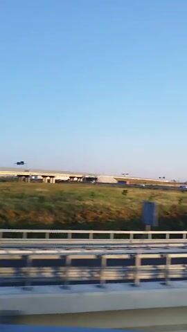 A video captured from a moving vehicle shows a highway with cars and a clear blue sky. In the foreground, a bridge railing is visible. The camera pans across a landscape with green fields and vegetation. A highway with multiple lanes is in the background, with several cars and trucks traveling on it. The sun appears to be low in the sky, casting long shadows. The overall impression is of a scenic drive on a highway.