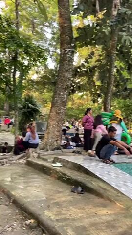 The video shows a park-like setting with many people present. In the foreground, there are concrete structures that appear to be part of a water feature or playground. Several people are sitting or squatting near these structures, some with their backs to the camera. Further back, more people are visible, some sitting on benches or on the ground. There are trees and lush greenery throughout the scene, suggesting an outdoor environment. In the middle ground, a group of people are gathered near what looks like a swimming pool. Some children are in the pool, and there are inflatable rings. The overall atmosphere appears relaxed and social, with people enjoying a day outdoors. The lighting suggests it might be late afternoon or early evening.