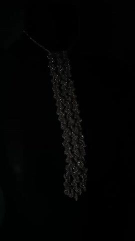 The video displays a close-up, high-angle shot of a sparkly, cascading necklace against a plain black background. The necklace appears to be made of numerous small, faceted beads or crystals that create a shimmering effect. It hangs from a delicate chain and drapes downwards in a textured, layered fashion, resembling a waterfall or fringe. The lighting focuses on the necklace, highlighting its intricate details and reflective qualities, while the background remains dark and featureless, ensuring the jewelry is the sole subject of attention. No people, location cues, text, or specific activity are visible in this media file. The time of day, weather, and any emotional context are not discernible from the provided image.