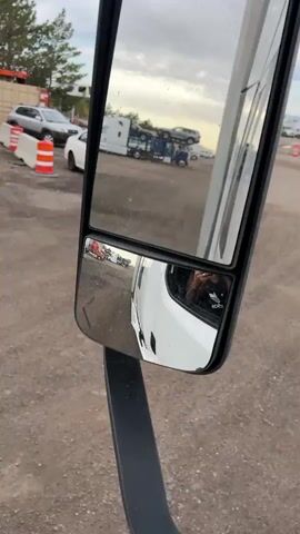 The media file shows a reflection in the side mirror of a white truck. The main mirror reflects a tow truck carrying two cars, a white trailer truck, and a white car. In the smaller, lower mirror, the reflection shows the side of the white truck the camera is in, with a person's hand holding a phone visible in the driver's side window. The scene appears to be a car lot or a yard where vehicles are stored or repaired. The ground is dirt and gravel, and there are other vehicles and structures in the background. The sky is cloudy, suggesting it might be late afternoon or early morning. There are no visible text elements in the media. The overall impression is a functional, outdoor setting related to vehicles.