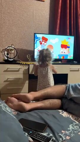 The video opens on a television screen displaying a cartoon with colorful animated characters. A young child with blonde curly hair, wearing a white tank top with a floral pattern, stands in front of the television, reaching towards it with their right hand. The child's back is to the camera. In the foreground, the bare legs and feet of an adult are visible, suggesting they are lying down. The adult's legs are crossed, and a smartphone and a remote control are on a surface in front of them. The environment appears to be a living room, with patterned wallpaper on the wall and a red curtain in the background. A dresser or cabinet is positioned beneath the television. The scene is likely set indoors in a home.