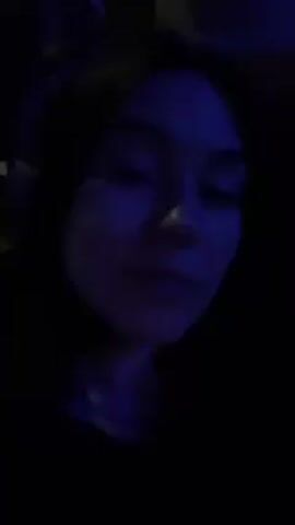 The video shows a person with shoulder-length dark hair wearing headphones, sitting in a car, possibly at night. The lighting is predominantly blue, making it difficult to discern specific facial features or details. The person is looking directly at the camera. In the background, another person can be briefly seen. The car appears to be moving, evidenced by the changing background.