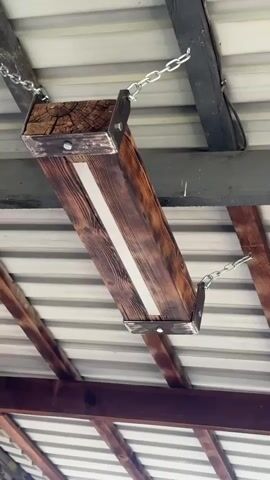 A close-up shot focuses on a rustic, wooden light fixture suspended from a metal roof and wooden beams by chains. The fixture is a rectangular wooden beam, stained dark brown with some lighter wood visible on its surface. A white LED light strip is embedded in the center of the beam, illuminating the area beneath. Metal brackets are attached to the ends of the wooden beam, and chains connect these brackets to the overhead structure. The lighting in the scene appears to be from the fixture itself and ambient light filtering from outside. The camera angle is looking up at the fixture from below, highlighting its construction and its integration with the ceiling.