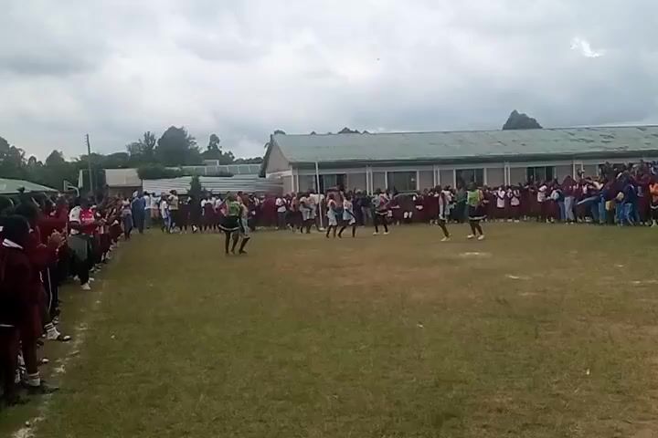 A wide shot shows a grassy field where a group of young women, dressed in matching green and black uniforms, are performing a dance routine. They are surrounded by a large crowd of spectators, mostly students, who are cheering and clapping. Some of the students are wearing maroon school uniforms while others are in casual clothes. The background features a long, single-story building with a light-colored roof, likely a school building. The sky is overcast, suggesting a cloudy day. The overall scene appears to be a school event or performance on a sports field.