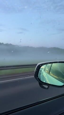 The video shows the interior of a car driving on a highway. A side view mirror is visible on the left side of the frame, reflecting the car's interior and the outside scenery. Raindrops are visible on the car's windows. The road is wet and visibility is reduced due to fog or mist. The sky is overcast and appears to be either dawn or dusk. In the foreground, a car dashboard with a touch screen display is visible. The display shows song titles and other audio information in Japanese. The car appears to be moving at a steady speed. The overall impression is of a journey through a misty, perhaps rainy, landscape.