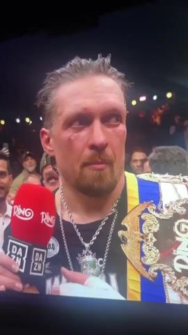 A close-up shot shows Oleksandr Usyk, a Ukrainian professional boxer, speaking into a red Ring and DAZN microphone. He has a bruised face, a wet, slicked-back hairstyle, and a trimmed beard. He wears a black t-shirt, a silver necklace with a crown pendant, and a Ring belt with a blue and yellow Ukrainian flag-colored stripe. The background shows a crowd of people, some holding up phones, and bright lights. Usyk appears to be giving a post-fight interview, referencing people in bomb shelters.
