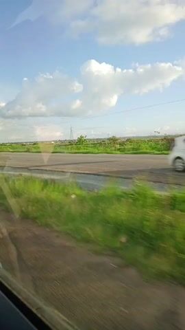 A car is traveling down a road on a sunny day with a blue sky and scattered white clouds. Power lines and towers are visible in the distance. Cars pass in the opposite direction. Green grass grows along the side of the road. The view is from inside the car, looking out the window.