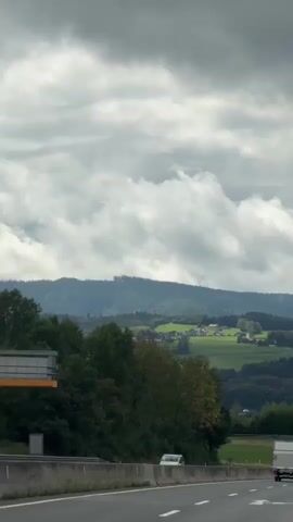 The video captures a view from a vehicle traveling on a highway. The highway has multiple lanes with white arrows painted on the road indicating the direction of travel. There are other vehicles on the highway, including cars and trucks. The landscape outside the highway features green hills, trees, and some buildings in the distance. The sky is cloudy. A blue road sign is visible, indicating "St. Georgen im Attergau". The scene appears to be a typical highway journey through a rural or semi-rural area.