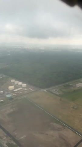 The video shows an aerial view from an airplane window, looking down towards the ground. The landscape is a mix of green forest, open fields, and infrastructure including roads and a facility with several cylindrical storage tanks. The visibility is limited due to a thick layer of clouds or fog. As the video progresses, the wing of a Ryanair airplane becomes visible in the frame, obscuring a portion of the ground view.
