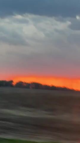 The video shows a sunset view from a moving vehicle, likely a car. The sun is setting over a field, creating an orange and red glow in the sky. There are trees silhouetted against the bright horizon. The sky has a mix of dark, cloudy areas and patches of pink and red from the sunset. There's a noticeable lens flare in the lower part of the frame. The camera movement suggests the car is traveling at a moderate speed.