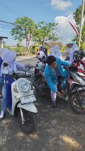 A group of young people, some wearing white uniforms and headscarves, and one in a blue blazer and white pants, are gathered around several parked motorcycles on a sunny day. The person in the blue blazer is interacting with a red motorcycle, appearing to adjust something. In the background, there are trees, a building with a thatched roof, and Indonesian flags are visible, suggesting a possible celebration or event. The ground is a mix of dirt and pavement, with some rocks and vegetation. The motorcycles appear to be the primary mode of transportation for the individuals present.