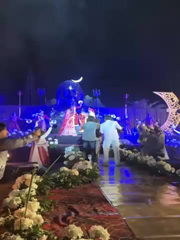 The video captures an outdoor wedding scene at night, with a large Shiva statue looming behind the stage. A bride in a red lehenga and a groom in a white sherwani stand on the stage, flanked by dancers also in red outfits. Several men, likely photographers, are taking pictures of the couple. The pathway leading to the stage is decorated with white flowers. Blue stage lighting illuminates the scene, creating a dramatic and festive atmosphere.
