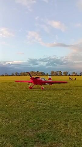 A red aerobatic airplane is parked on a grassy field. The sky is cloudy with patches of blue. Trees and distant buildings can be seen in the background.