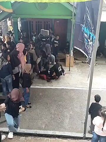 A group of people are gathered in front of a house, some standing and some sitting on the ground. A sound system with speakers and a mixer is set up. A large black banner with white lettering is displayed on the right side of the frame. Some people are looking at their phones, while others appear to be interacting with each other. A few children are visible on the right side of the frame. The scene suggests an informal gathering or event, possibly with music or a performance.
