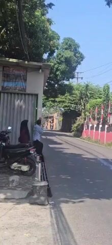 A short video shows a street scene on a sunny day. On the left side of the street, a building with a corrugated metal wall and a poster is visible. A red and black motorcycle is parked in front of the building. Two people, a woman in a hijab and a man in a blue shirt, stand near the motorcycle, facing the street.

The street itself is paved and appears to be a two-lane road. There are trees and vegetation along the sides of the road, and a few buildings can be seen in the distance. On the right side of the street, a red and white striped barrier or fence runs along the sidewalk. Several Indonesian flags are visible on poles along this barrier.

Several motorcycles are seen riding down the street. The first two are individuals riding them. The third motorcycle is carrying a large load of green leaves, which completely covers the back of the motorcycle and appears to be piled high. The video is taken from a slightly elevated position, looking down at the street.