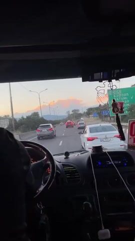This video provides an interior perspective from a vehicle traveling on a highway. The view is directed forward through the windshield, showing a road with multiple lanes and moderate traffic. The sky is visible above, displaying a sunset with illuminated clouds in shades of orange and pink. Several cars are driving in the same direction, with some appearing to be moving at a similar speed, suggesting a slight traffic jam or congestion. The dashboard of the vehicle is partially visible, including the steering wheel, instrument cluster, and the center console with some electronic devices. Hanging from the rearview mirror are religious ornaments, specifically a cross with praying hands and a small teddy bear charm. The environment appears to be rural or suburban, with trees and hills visible in the distance. The overall impression is a journey in a vehicle during twilight.