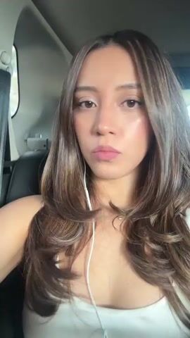 A young woman with long, layered brown hair, with blonde highlights, is sitting in the back seat of a car. She is wearing a white tank top and has earbuds in her ears. She looks at the camera and blinks. The interior of the car is visible, including a black leather seat and part of a headrest. The background shows a blurry outdoor scene with some trees and what appears to be a building or structure. The lighting suggests it is daytime.