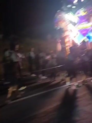 The video shows a nighttime street scene with a crowd of people. A brightly lit float or decoration, possibly a dragon, is visible on the right side of the frame. People are standing and walking along the side of the road, some appear to be watching the float. A few individuals are near a motorized vehicle, and others are gathered on a grassy embankment in the background. The lighting is dim, with the main illumination coming from the decorative structure. The atmosphere appears to be festive, possibly a parade or celebration.