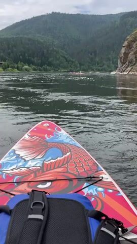 The video begins with a close-up of the front of a paddleboard adorned with a colorful koi fish design. A blue and black backpack is secured to the board. The camera then pans out to reveal a wide, fast-flowing river with choppy water. In the distance, on the river, a group of several red-colored rafts or boats are visible, moving downstream. The riverbanks are lined with lush green, densely forested mountains. The sky is overcast with clouds, suggesting a temperate climate. The overall impression is an outdoor activity, likely a rafting or paddleboarding excursion on a natural waterway surrounded by mountainous terrain.