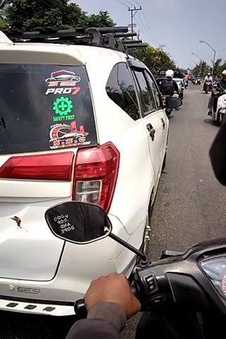 A first-person perspective video shows traffic on a paved road. The camera is mounted on a motorcycle, facing forward. To the left, a white car is visible, with stickers on its rear window. Several motorcycles are on the road, some with riders wearing helmets. The environment appears to be a roadside with trees and vegetation. The sky is visible and appears to be daytime with some clouds. The car to the left has a dented rear bumper.