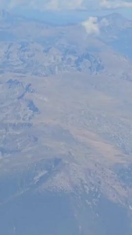 The video provides an aerial view of a mountainous landscape under a clear blue sky with scattered clouds. The foreground features the wing of an airplane. The mountains are rugged and appear to be arid or sparsely vegetated, with visible ridges and valleys. The sunlight illuminates the terrain, casting shadows and highlighting the topography. The clouds are puffy and white, adding depth to the sky. The airplane wing, a prominent object in the frame, is light gray with some black markings and a yellow striped caution area. The perspective suggests the camera is looking out of an airplane window as it flies over the mountains.