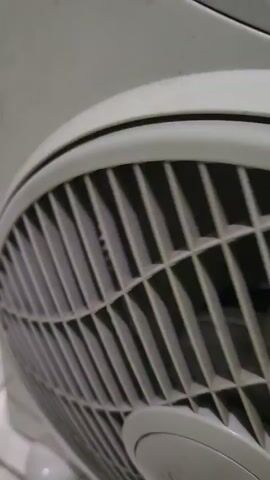 A close-up, slightly shaky video shows the grille of a white electric fan. The fan's blades are visible behind the grille and appear to be rotating. The fan is light-colored, possibly white or off-white, and has a textured surface. The background is out of focus, but appears to be a light-colored wall or surface.