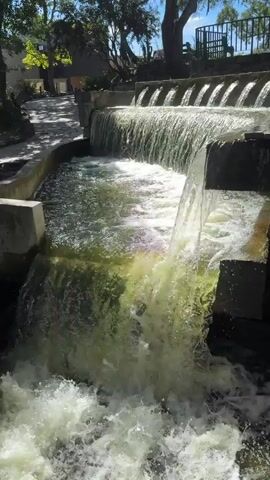 The video depicts a stepped concrete waterfall. The water flows down in stages, creating white water at the bottom. The water appears slightly green in color. The environment is a park-like setting, with trees visible in the background and a paved walkway to the left of the waterfall. A building is visible in the distance. There are people on a bench in the background behind a fence. The camera remains stationary throughout the video.