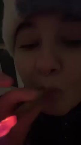 A close-up video shows a person wearing a white beanie exhaling vapor, possibly from an electronic cigarette or vaping device. The person has a nose ring and is wearing a dark jacket. The background is dark and indistinct.