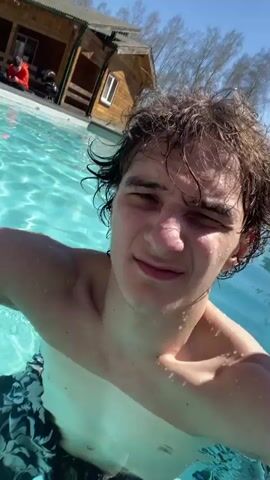 A young man with dark hair and pale skin is swimming in a pool. He is wearing dark swim trunks. The water is clear and blue, with sunlight creating patterns on the surface. The man appears to be holding a camera, filming himself as he submerges his head underwater. In the background, there is a small wooden building, a white car, and a few people sitting near the pool. The sky is clear and sunny. The camera is close to the man's face, capturing his expressions as he interacts with the water.