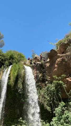 A group of people are rappelling down a rocky cliff next to a waterfall on a sunny day. The individuals are wearing helmets and harnesses and are secured by ropes. One person is actively descending the cliff, while others are positioned at the top, assisting or observing. The environment is lush with green vegetation and a clear blue sky.
