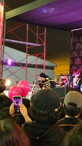 The video shows a crowd of people at a live concert. The band, dressed in black and gold uniforms, performs on a stage illuminated by purple and white lights. A banner with the name "Eno" and "Mariana" is visible to the left of the stage. People in the crowd are holding up their phones to record the performance, and pink balloons bob above the heads of the audience. The stage is covered by a purple awning, and scaffolding is visible behind the band.