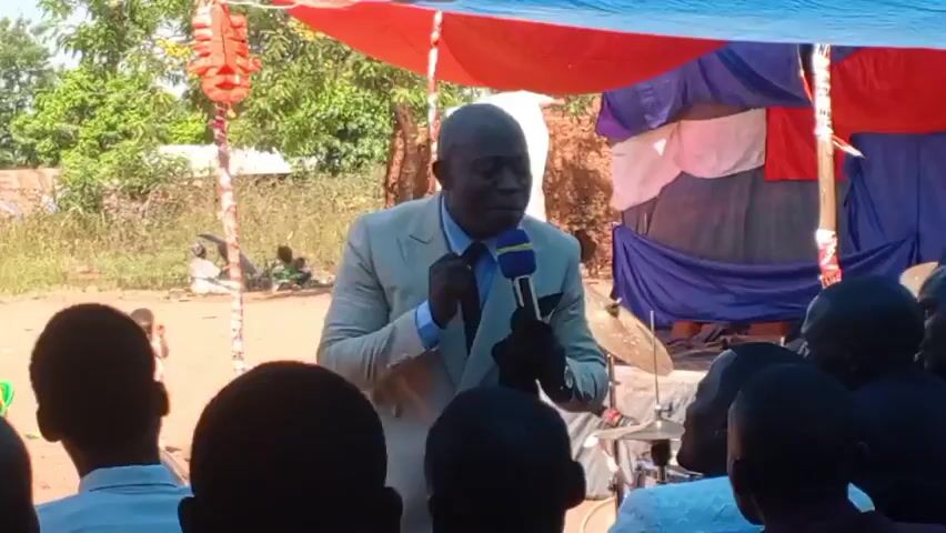 A man in a light-colored suit, with a microphone, addresses a crowd of people. The setting appears to be outdoors, under a makeshift canopy constructed of tarps. People are seated in chairs and on the ground. A sound system is visible. A man in a white shirt films the speaker with his phone. The environment consists of trees and buildings in the background.