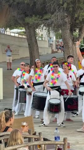 In this vertical video clip, a group of individuals are seen playing drums as part of an outdoor performance. They are wearing matching white outfits, including loose-fitting pants and t-shirts, and colorful leis around their necks. The drummers are positioned in a line, with some holding large bass drums and others smaller drums. The performance appears to be taking place on a wooden boardwalk, possibly at a beachside venue.

In the background, trees with sparse foliage provide shade, and a pathway lined with plants is visible. Further back, a white building with the word "mar" faintly visible on it suggests a coastal location. Several spectators are observing the performance from a distance, some seated and others standing. The overall atmosphere seems festive and energetic, with the drummers engaged in their music and the audience appearing to enjoy the spectacle. One individual, facing away from the camera, is gesturing with their arm, possibly directing the performance.

The lighting suggests it is daytime, likely late afternoon or early evening, given the soft, diffused light filtering through the trees. There are no strong shadows, and the colors are natural, indicating pleasant weather. On the drums, the word "BATAMBADA TERRAS" is visible, likely indicating the name of the drumming group. A water bottle is placed on the boardwalk in the foreground.