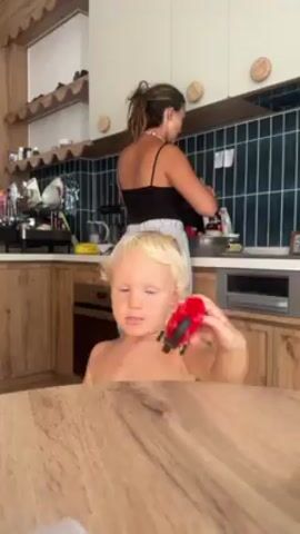 The video shows a young child playing with a red toy motorcycle on a wooden table. The child is fair-skinned, with blond hair and light-colored eyes. They are partially visible from the chest up. In the background, a woman is standing at a kitchen counter, facing away from the camera. She is wearing a black tank top and light-colored, wide-legged pants. She has her hair pulled back in a ponytail and appears to be preparing food. The kitchen has light-colored cabinets, dark teal tiled backsplash, and wooden countertops. There are various kitchen items on the shelves and countertops. The lighting appears to be natural daylight.