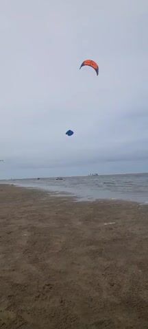 The video opens with a wide shot of a beach and the ocean under an overcast sky. In the foreground, the sandy beach stretches towards the water's edge. The ocean appears calm with small waves lapping at the shore. In the distance, several people can be seen in the water, some of whom are participating in kitesurfing. Two kites are visible in the sky: a larger orange and blue kite in the upper right portion of the frame, and a smaller blue kite further to the left and lower down. The camera perspective is low, pointing upwards towards the sky and the kites. The overall scene depicts a peaceful day at the beach with recreational activities taking place on the water.