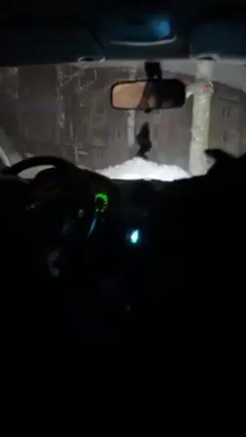 A person is sitting in the driver's seat of a car at night. The interior of the car is dimly lit, with the dashboard gauges and radio illuminated. Outside, the car is facing what appears to be a snowy landscape, illuminated by the car's headlights. Music is playing, with a distinct bass tone. The car is stationary.
