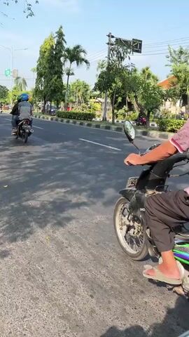 The video shows a street scene in what appears to be a tropical or subtropical climate. The perspective is from someone riding a motorcycle, looking forward and slightly to the right.

**People:**
*   The person filming is riding a motorcycle, wearing dark pants and a maroon and white patterned shirt. Their right hand is visible on the handlebars, and they are wearing a sandal on their right foot.
*   Another motorcycle is visible ahead, moving in the same direction. It carries two people: a driver wearing a blue helmet and a passenger behind them.

**Objects:**
*   Two motorcycles are the primary objects.
*   Street signs and a traffic light are visible in the background.
*   Trees, including palm trees and other lush vegetation, line the street.

**Actions:**
*   The motorcycles are in motion, traveling down a paved road.
*   The sun is casting distinct shadows from the trees onto the road, indicating a sunny day.

**Environment:**
*   The scene is an urban or suburban street with a clear blue sky overhead.
*   The road is asphalt and appears to be in good condition.
*   There is a median or planted area between lanes of traffic, with trees and shrubs.
*   Buildings or structures are visible in the distance, along with more greenery.

**Interactions:**
*   The two motorcycles are traveling in the same lane, with the filming motorcycle behind the lead motorcycle. There is no direct interaction visible, only proximity and shared direction of travel.

**Overall Impression:**
The video captures a common mode of transportation and everyday life in a warm climate, focusing on the perspective of a motorcyclist navigating a street with other traffic and abundant greenery.