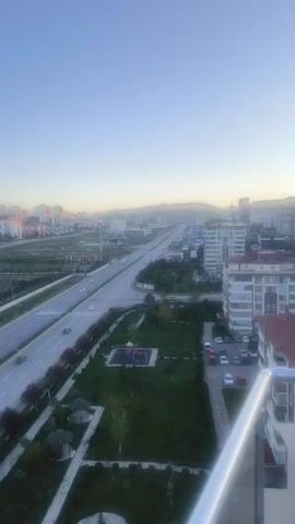 The video presents a high-angle, sweeping view of a city landscape during either sunrise or sunset, evidenced by the low sun casting golden reflections on distant buildings. A wide, multi-lane road with cars traveling in both directions dominates the foreground and midground, flanked by green spaces, trees, and small gazebos. To the right of the road, there's a park area, and further left, residential buildings and undeveloped land with planted trees. As the camera pans left, a tennis court and a soccer field become visible, along with an area showing construction equipment (tractors, trailers) and a small building with a red roof. In the background, numerous modern residential buildings extend towards mountains. The sky is clear and blue. No people are clearly visible, and any text on a small sign near the road is illegible. The scene depicts a modern urban or suburban environment, consistent with Etimesgut, Turkey.