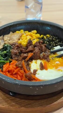 A close-up shot shows a stone bowl filled with bibimbap, sitting on a wooden tray. The bibimbap includes beef, corn, shredded carrots, spinach, mushrooms, seaweed, and a fried egg. Chopsticks are used to break the yolk of the fried egg. A bottle of water stands in the background.