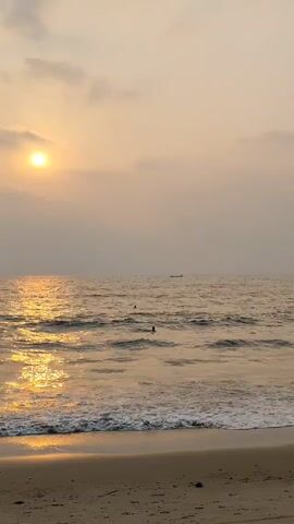 The scene displays a beach at sunset. The sun is visible through a cloudy sky, casting a golden reflection on the ocean surface. Gentle waves are rolling onto the sandy beach. Several figures are seen swimming in the ocean. A distant ship can be seen on the horizon.